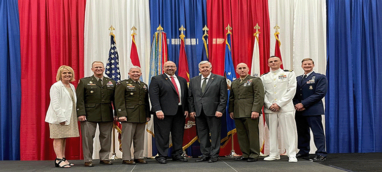 A photo of military, state, and community leaders standing in front of a red, white, and blue background and flags. 