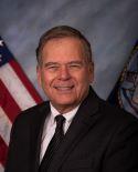 Admiral Charles Williams, USN, Retired wearing a black suit and tie in front of two flags.